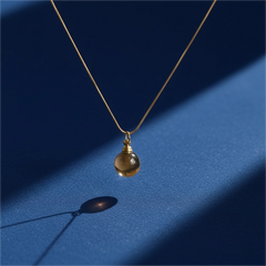 Yellow agate urn necklace on gold chain, resting on blue background under natural sunlight, symbol of warmth and calm.